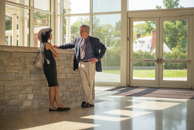 A man and woman standing in a well-lit lobby make casual conversation in front of a stone brick wall